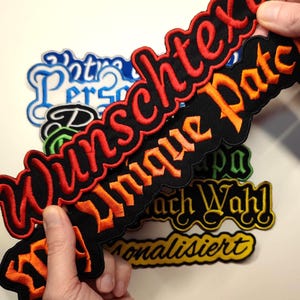 Op de afbeelding: Geborduurde patches in verschillende kleuren en lettertypen. De patches bevatten teksten zoals "Wunschtext" in rood, "Unique Pate" in oranje en andere in blauw, groen en geel. De patches worden door een persoon vastgehouden.