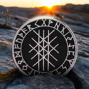 Op de afbeelding: Zwarte geborduurde patch met een wit Vegvisir-symbool omringd door een cirkel van runen. De patch ligt op een rotsachtig oppervlak.