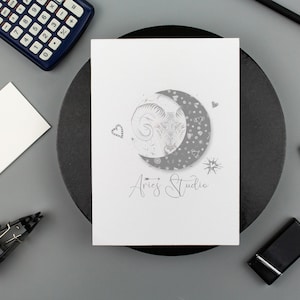 Könnte beinhalten: Eine weiße rechteckige Karte mit einer kreisförmigen Grafik eines Widderkopfes in einer Mondsichel, umgeben von kleinen Stern- und Herzsymbolen. Unter der Grafik ist der Text Aries Studio in einer Schreibschrift aufgedruckt.