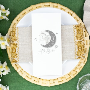 Könnte beinhalten: Eine weiße, rechteckige Menükarte mit einer grauen Grafik eines Widderkopfes in einem Halbmond und dem Text Aries Studio. Die Karte liegt auf einer beigen Leinen-Serviette mit goldener Blumenstickerei auf einem runden Rattan-Platzset.