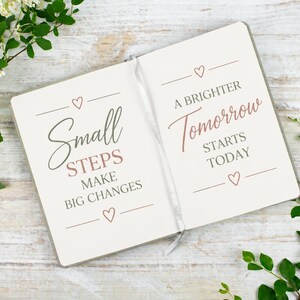 Puede incluir: Un cuaderno abierto de páginas blancas con texto en gris y oro rosa. La página izquierda dice Small STEPS MAKE BIG CHANGES con corazones. La página derecha dice A BRIGHTER Tomorrow STARTS TODAY con corazones y un marcapáginas de cinta.