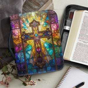 May include: A colorful Bible cover featuring a stained glass design with a cross at the center. The cover is predominantly purple, yellow, and blue, with a dark brown border. A Bible is partially visible inside a black case, along with a pen and notepad.