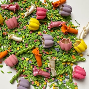 May include: A colorful assortment of edible sugar decorations, including pink, yellow, orange, and purple flower shapes, green stems, and tiny flowers, scattered on a bed of green sprinkles. These are likely cake or cupcake decorations.