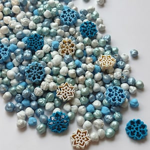 Puede incluir: Primer plano de un montón de chispas perladas blancas, azules y plateadas con decoraciones en forma de copo de nieve. Los copos de nieve son en tonos azules y blanquecinos. Las chispas están esparcidas sobre una superficie blanca.