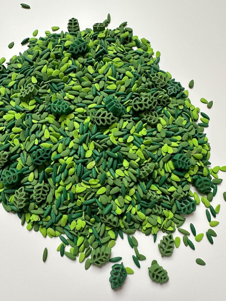 May include: A collection of small, green decorative sprinkles in various shades of lime, forest, and emerald green. The mix includes numerous tiny, flat, oval-shaped pieces alongside several larger, detailed, three-dimensional leaf shapes.