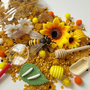 Puede incluir: Un recipiente sensorial con pasta amarilla, girasoles artificiales, una figura de abeja y una lupa. El recipiente también incluye pequeños elementos decorativos como pompones, un cucharón de miel de madera y una hoja con formas blancas. Los elementos están dispuestos sobre una superficie blanca.