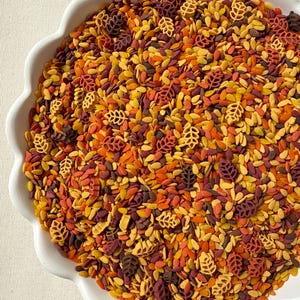 May include: A white scalloped bowl filled with a mixture of dry pasta shapes in autumn colours. The blend includes small yellow, orange, and dark red grain-like pieces mixed with larger, detailed pasta shapes resembling various types of leaves.
