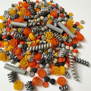Può includere: Un assortimento colorato di forme di pasta, tra cui ruote, spirali e tubi, in tonalità di giallo, arancione, grigio e nero. La pasta è sparsa su una superficie bianca, creando un'esposizione vibrante e strutturata.