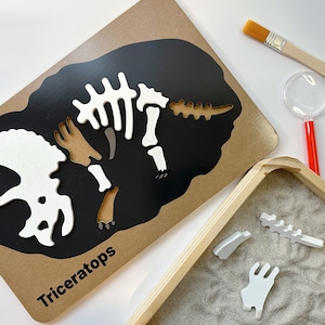 Pode incluir: Um quebra-cabeça de esqueleto de dinossauro de madeira com uma silhueta preta de Triceratops e peças de quebra-cabeça brancas. As peças do quebra-cabeça estão espalhadas em uma bandeja de areia branca. O conjunto também inclui uma lupa e um pincel.
