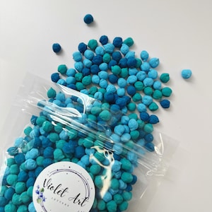 May include: A clear plastic bag filled with small, round, blue and teal paper confetti. The bag is partially open, and some of the confetti has spilled out. The bag has a white sticker with the text "Violet Art Letters" on it.