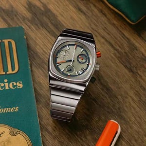 Pode incluir: Um relógio de pulso prateado com mostrador quadrado e pulseira de metal. O mostrador do relógio é verde claro com detalhes em laranja e azul. O relógio é exibido em uma superfície de madeira ao lado de um livro.