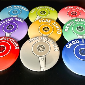 May include: Nine round buttons with different colors and names of the members of the K-pop group Twice. Each button features a stylized image of a microphone with the group's logo.