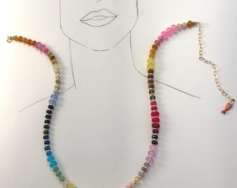 Puede incluir: Un collar de cuentas multicolor que presenta un degradado de pequeñas cuentas redondas y translúcidas en tonos rosa, amarillo, marrón, azul y verde. El collar incluye espaciadores dorados y una cadena extensora dorada con un pequeño dije de pez naranja.