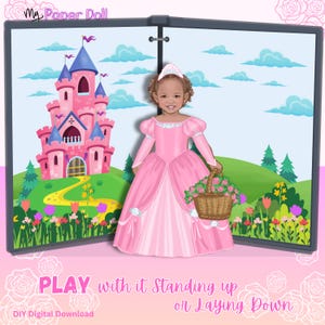 May include: A My Paper Doll digital download featuring a paper doll character wearing a pink princess dress and tiara, holding a basket of flowers. The background shows a pink castle, green hills, and flowers. Text reads: PLAY with it Standing up or Laying Down.