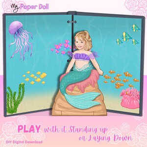 May include: A My Paper Doll digital download featuring a mermaid character with blonde hair and a teal tail sitting on a rock. The background shows an underwater scene with a purple jellyfish, green seaweed, a pink anemone, and various small fish.