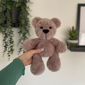 Op de afbeelding: Een pluche teddybeer in een zachte, gedempte mauve kleur. De beer heeft een zwart gestikte neus en een eenvoudig, innemend ontwerp. De vacht ziet er pluizig en getextureerd uit, wat duidt op een knuffel. Een klassiek kinderspeelgoed.