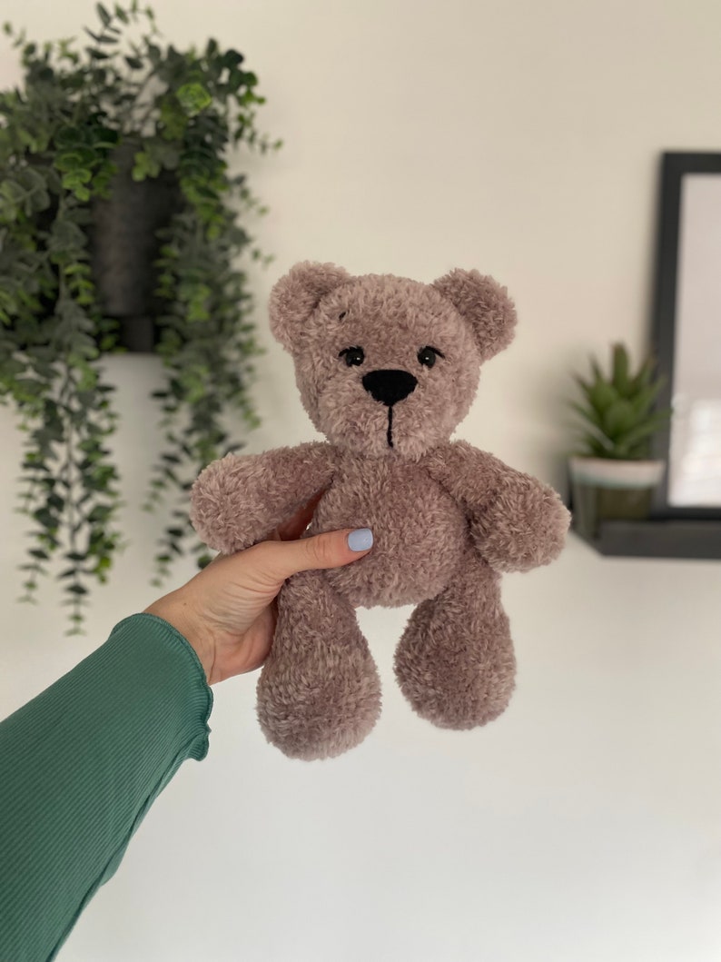 Op de afbeelding: Een pluche teddybeer in een zachte, gedempte mauve kleur. De beer heeft een zwart gestikte neus en een eenvoudig, innemend ontwerp. De vacht ziet er pluizig en getextureerd uit, wat duidt op een knuffel. Een klassiek kinderspeelgoed.