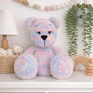 Op de afbeelding: Een grote, zachte teddybeer in pastelkleuren met een zwarte neus en ogen. De beer is gemaakt van zacht garen in roze, blauwe, gele en groene tinten. Hij zit op een wit oppervlak. De beer is een kinderspeelgoed.