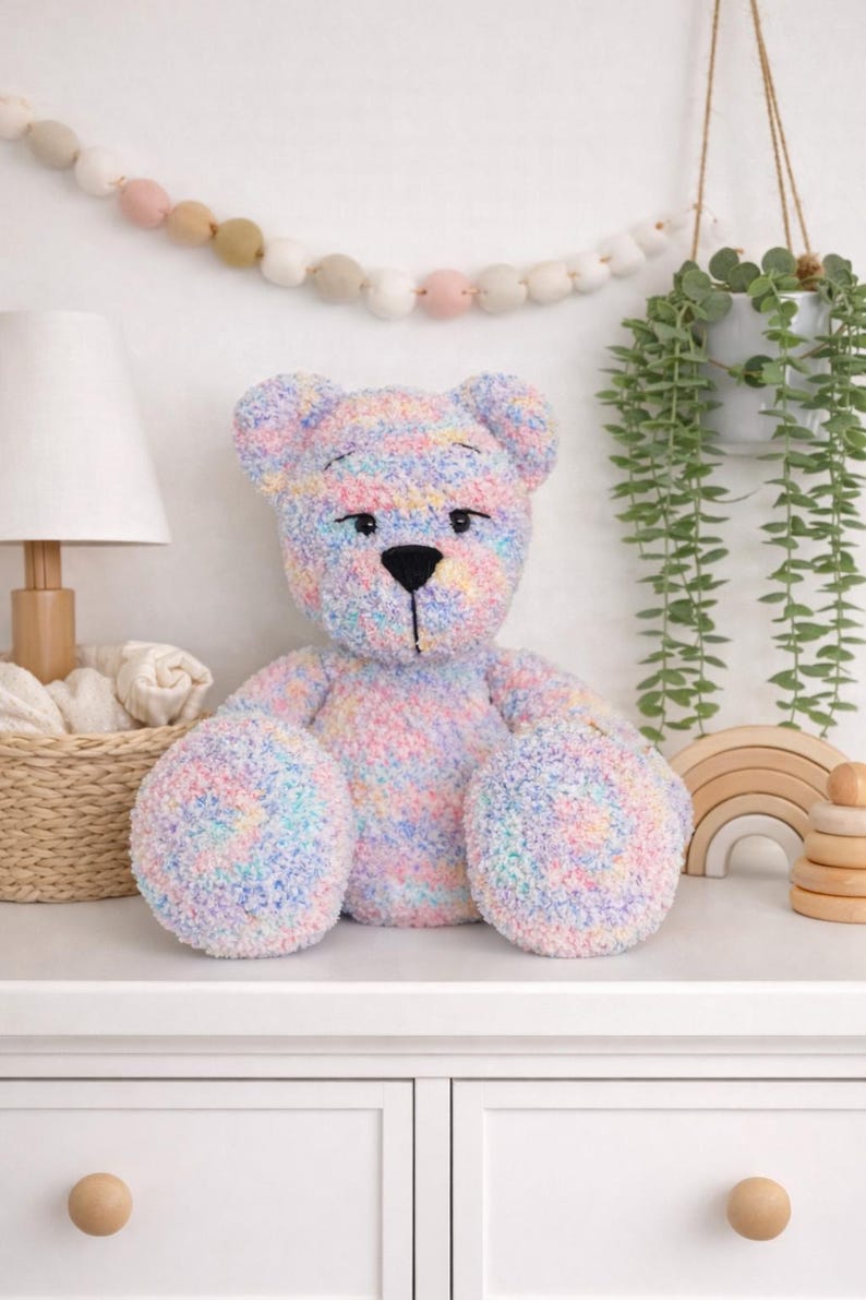 Op de afbeelding: Een grote, zachte teddybeer in pastelkleuren met een zwarte neus en ogen. De beer is gemaakt van zacht garen in roze, blauwe, gele en groene tinten. Hij zit op een wit oppervlak. De beer is een kinderspeelgoed.