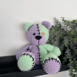 May include: A crocheted teddy bear with a green and purple patchwork design. The bear has black eyes and a brown button nose. The bear is sitting on a shelf.