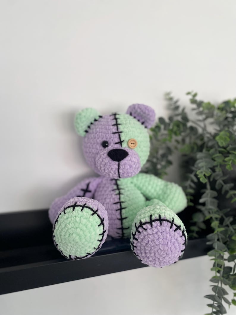 May include: A crocheted teddy bear with a green and purple patchwork design. The bear has black eyes and a brown button nose. The bear is sitting on a shelf.