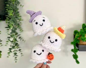 May include: Three crocheted ghost ornaments with different Halloween-themed hats. The top ghost wears a purple witch hat, the middle ghost wears a yellow and orange candy corn hat, and the bottom ghost wears a small orange pumpkin.
