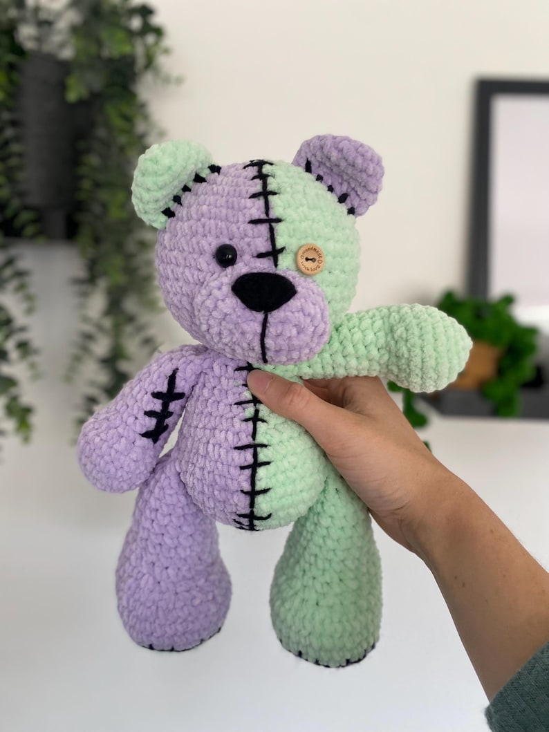 May include: A crocheted plush toy bear with a green and purple body, black stitching, and a wooden button eye.