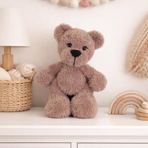 Op de afbeelding: Een zachte, lichtbruine teddybeer met een zwarte neus en ogen staat op een wit oppervlak. De beer wordt omringd door een decoratieve slinger, een lamp, een rieten mand en houten speelgoed. De algehele esthetiek is zacht en uitnodigend.