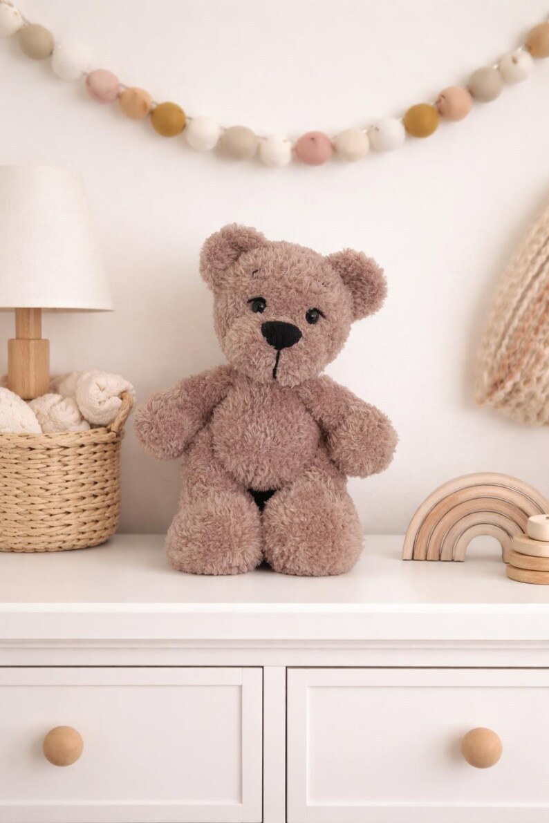 Op de afbeelding: Een zachte, lichtbruine teddybeer met een zwarte neus en ogen staat op een wit oppervlak. De beer wordt omringd door een decoratieve slinger, een lamp, een rieten mand en houten speelgoed. De algehele esthetiek is zacht en uitnodigend.