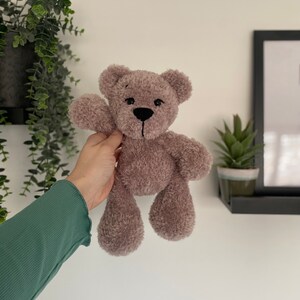 Op de afbeelding: Een zachte, taupekleurige teddybeer met een zwart gestikte neus en mond. De beer heeft een zachte, fuzzy textuur en zwaait met &eacute;&eacute;n arm. De beer is ongeveer 20 cm hoog.