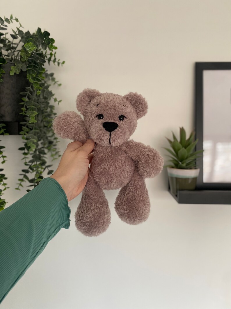 Op de afbeelding: Een zachte, taupekleurige teddybeer met een zwart gestikte neus en mond. De beer heeft een zachte, fuzzy textuur en zwaait met &eacute;&eacute;n arm. De beer is ongeveer 20 cm hoog.