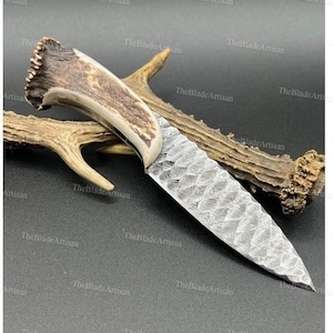 Puede incluir: Un cuchillo hecho a mano con una hoja de acero martillada texturizada y un mango de asta de ciervo. El cuchillo está apoyado en un trozo de asta de ciervo.