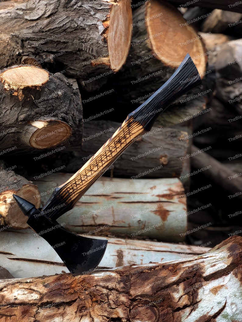 Pu&ograve; includere: Un'ascia nera con manico in legno. Il manico &egrave; decorato con un motivo geometrico e ha una macchia marrone scuro. L'ascia &egrave; appoggiata su una pila di tronchi.