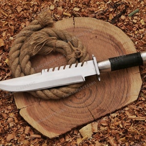 May include: A silver knife with a black handle and a serrated blade. The knife is lying on a wooden surface with a rope.
