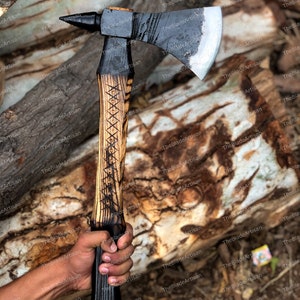 Pu&ograve; includere: Un'ascia nera e argentata con un manico di legno. Il manico &egrave; decorato con un motivo intagliato. L'ascia &egrave; tenuta in una mano.