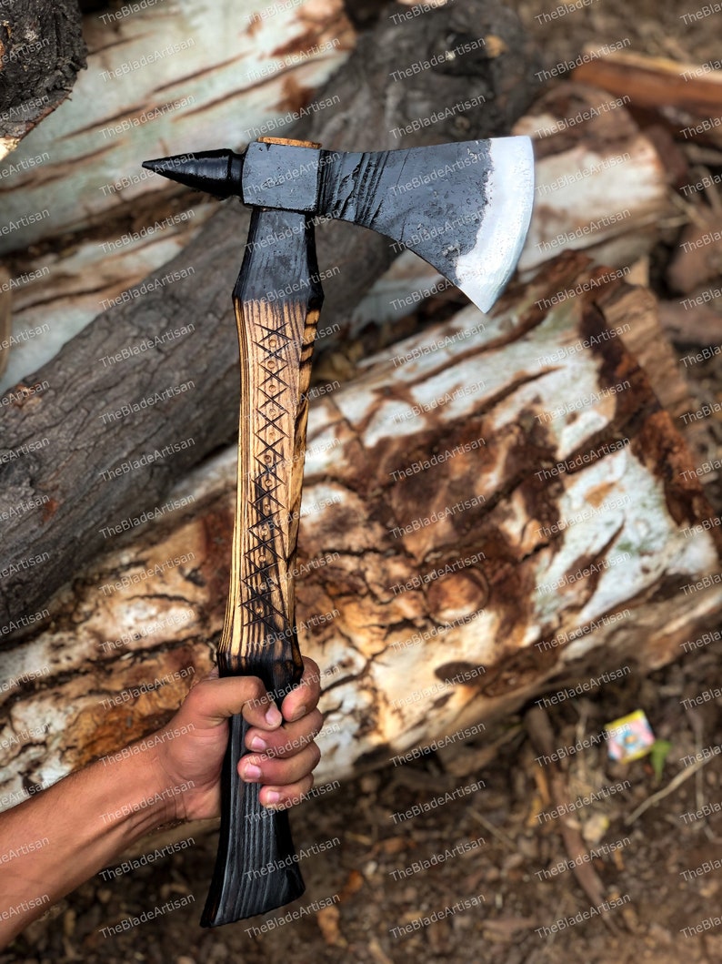 Pu&ograve; includere: Un'ascia nera e argentata con un manico di legno. Il manico &egrave; decorato con un motivo intagliato. L'ascia &egrave; tenuta in una mano.