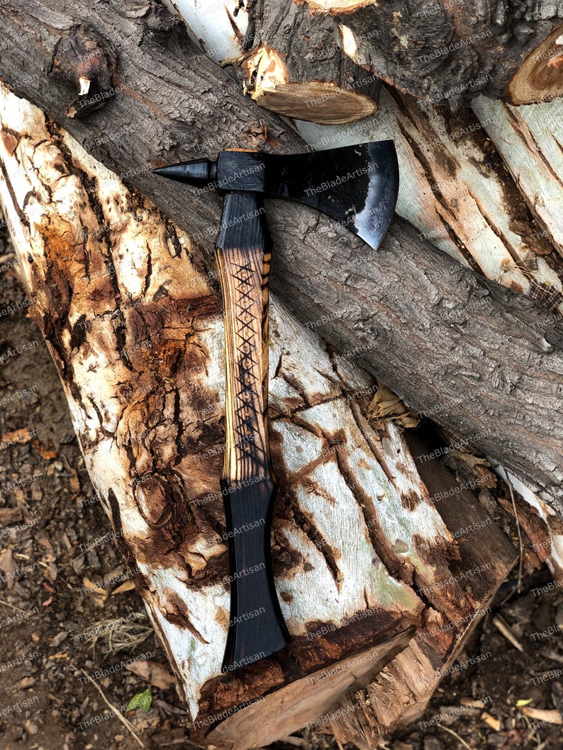 Pu&ograve; includere: Un'ascia nera con un manico di legno. Il manico &egrave; decorato con un motivo intagliato. L'ascia &egrave; appoggiata su una pila di tronchi.
