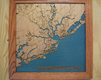 May include: A wooden map of Charleston, South Carolina, framed in a natural wood frame. The map is laser-cut and shows the city's streets and waterways. The background is a teal blue color. The text "CHARLESTON SC" is engraved below the map.