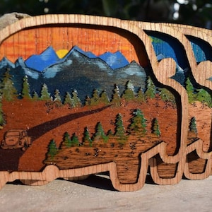 May include: Three wooden cutouts of a vintage car driving through a mountain landscape. The car is brown and the landscape is painted with a sunset, blue mountains, and green trees.