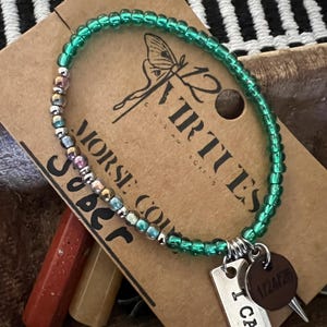May include: Green beaded bracelet with a silver charm that says "I Can" and a brown charm with the text "12 Virtues". The bracelet also has colorful beads that spell out "Sober" and "Morse Code".