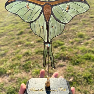 May include: A stained glass luna moth sculpture with pale green wings and amber accents, mounted on a black rectangular wooden base. The base features the gold-colored text "Trust the Unseen Path" and is decorated with small beads on the metal support rod.