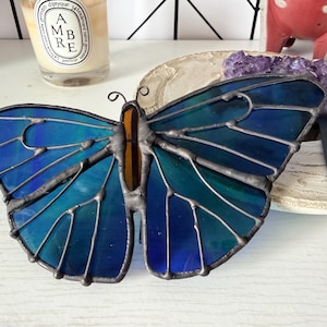 May include: A stained glass butterfly with blue and teal wings and an orange body. The butterfly is made with lead came and sits on a white surface. The wings have a detailed pattern.