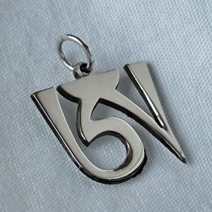 Può includere: Un ciondolo d'argento a forma di simbolo Om stilizzato. Il ciondolo ha un anello nella parte superiore per una catena ed è realizzato in metallo lucido. Il design presenta il tradizionale simbolo buddista tibetano.