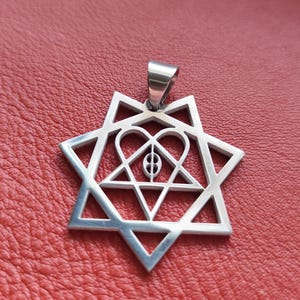 Può includere: Ciondolo color argento con design a Stella di David, con un cuore e forme geometriche al centro. Il ciondolo ha un anello per una catena ed è posto su uno sfondo rosso.
