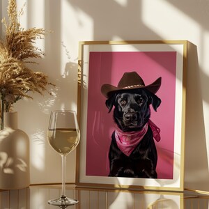 K&ouml;nnte beinhalten: Ein gerahmter Druck eines schwarzen Hundes, der einen braunen Cowboyhut und ein rosafarbenes Bandana vor einem rosafarbenen Hintergrund tr&auml;gt.