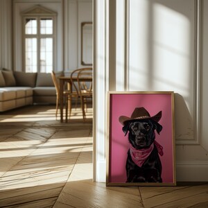 K&ouml;nnte beinhalten: Ein gerahmter Druck eines schwarzen Hundes, der einen braunen Cowboyhut und ein rosafarbenes Bandana vor einem rosafarbenen Hintergrund tr&auml;gt.