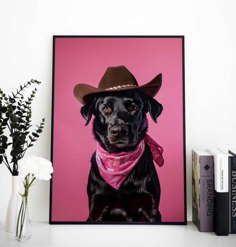K&ouml;nnte beinhalten: Ein schwarzer Labrador Retriever mit einem braunen Cowboyhut und einem rosafarbenen Bandana vor einem rosafarbenen Hintergrund.