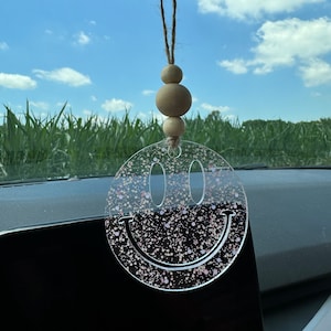 May include: A clear acrylic smiley face car charm with pink and gold glitter. The charm is hanging from a rearview mirror with a brown string and wooden beads.