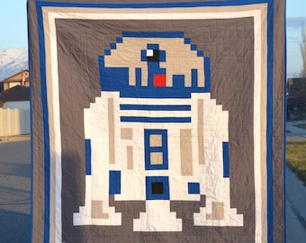 May include: A gray and white quilt with a pixelated image of R2-D2 from Star Wars. The quilt is framed with a blue and white border.