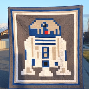 May include: A gray and white quilt with a pixelated image of R2-D2 from Star Wars. The quilt is framed with a blue and white border.